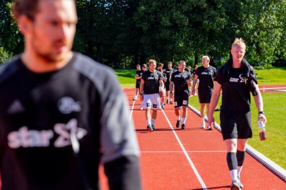Eine Gruppe von Personen in schwarzer Sportkleidung läuft auf einer roten Laufbahn im Freien, mit Bäumen und grünem Gras im Hintergrund. Eine Person im Vordergrund ist unscharf.