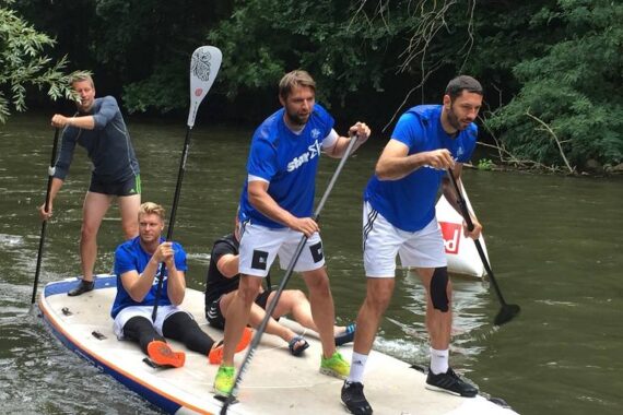 Vier Männer in blauen Hemden und weißen Shorts fahren gemeinsam Stand-up-Paddleboarding auf einem Fluss, zwei stehen und paddeln, während zwei vorne auf dem Brett sitzen. Im Hintergrund sind Bäume zu sehen.