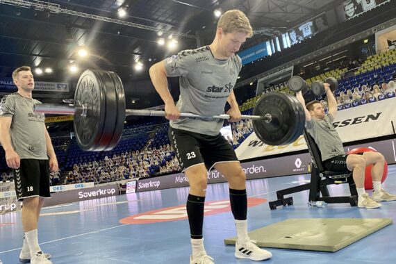 Drei Athleten in passenden grauen Hemden und schwarzen Shorts stemmen Hanteln auf einem Hallensportplatz, im Hintergrund sind Stadionplätze und Zuschauer zu sehen.