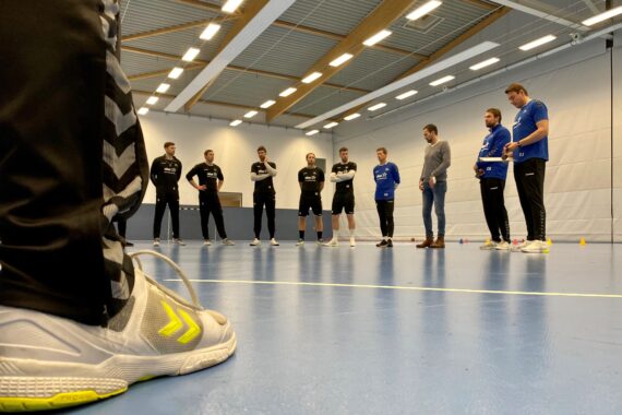 Eine Gruppe von Männern in Sportkleidung steht in einem Halbkreis auf einem Hallenplatz und hört einem Trainer zu. Der Fokus liegt auf einer Nahaufnahme eines Fußes in einem weißen Turnschuh im Vordergrund. Die Turnhalle hat hohe Decken und helle Lichter.