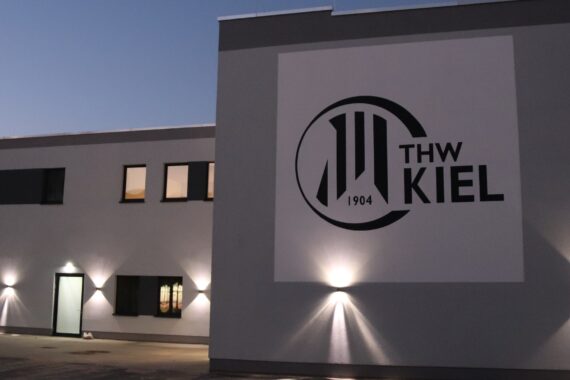 Ein modernes Gebäude in der Abenddämmerung mit dem Logo und dem Schriftzug THW KIEL 1904 an der Wand, beleuchtet von Außenleuchten.
