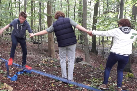 Drei Personen balancieren auf einer Slackline in einem Wald, wobei sie sich an den Händen abstützen. Der Boden ist mit Blättern bedeckt, und Bäume umgeben sie. Sie scheinen sich darauf zu konzentrieren, ihr Gleichgewicht zu halten.