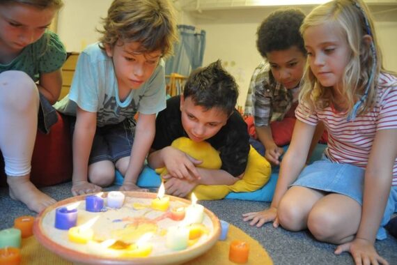 Vier Kinder sitzen dicht gedrängt um ein flaches Tablett mit Sand und bunten, brennenden Kerzen und beobachten aufmerksam die Flammen in einem gemütlichen Raum.