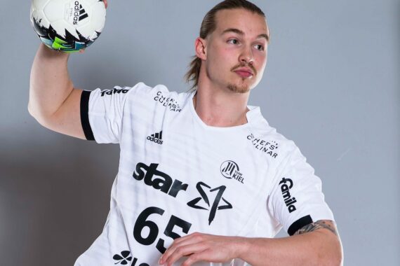 Ein männlicher Handballspieler in einem weißen Trikot mit der Nummer 65 und Sponsorenlogos bereitet sich darauf vor, einen Ball zu werfen, und schaut konzentriert. Er hat hellbraunes, zurückgebundenes Haar und einen Schnurrbart. Der Hintergrund ist einfarbig grau.