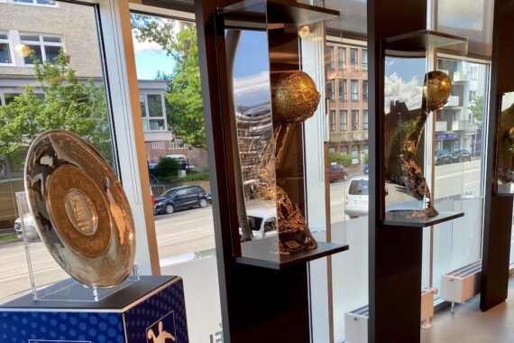 Ein großes silbernes Fußballmeisterschaftsschild und zwei goldene Weltmeisterschaftstrophäen sind in Glasvitrinen an einem Fenster ausgestellt, aus dem man an einem sonnigen Tag Autos und Gebäude sehen kann.