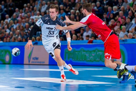 Zwei männliche Handballspieler kämpfen auf einem Hallenplatz um den Ball. Der eine, in einer weiß-schwarzen Uniform, greift nach dem Ball, während der andere, in Rot, versucht, ihn zu blockieren. Die Zuschauer füllen die Tribünen im Hintergrund.