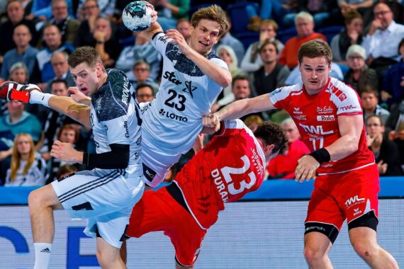 Drei Handballspieler in weißen und roten Trikots kämpfen während eines Spiels intensiv um den Ball, während im Hintergrund ein Publikum zusieht. Die Szene ist actionreich und körperbetont.