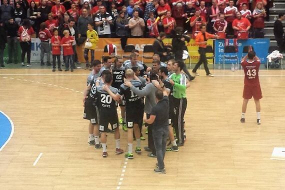 Eine Handballmannschaft in schwarzen und grauen Trikots versammelt sich auf dem Spielfeld, umgeben von Fans in roten Hemden, die auf der Tribüne jubeln. Ein Spieler in einem roten Trikot steht allein daneben.