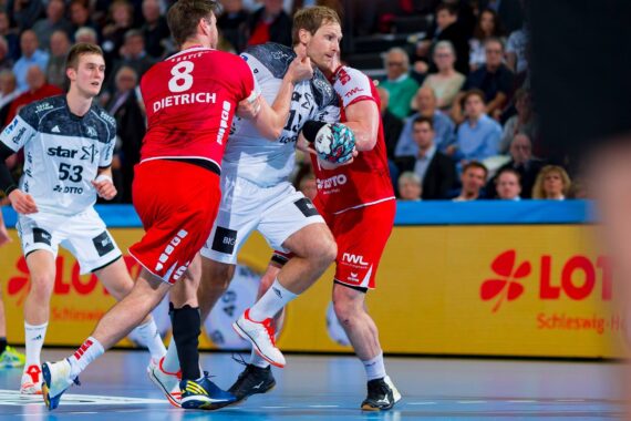 Zwei Handballspieler in rot-weißen Trikots sind in Aktion, wobei der eine Spieler den anderen mit dem Arm blockiert. Ein dritter Spieler schaut in der Nähe zu, und im Hintergrund ist eine Menschenmenge zu sehen. Die Szene findet in einer Halle auf einem blauen Spielfeld statt.