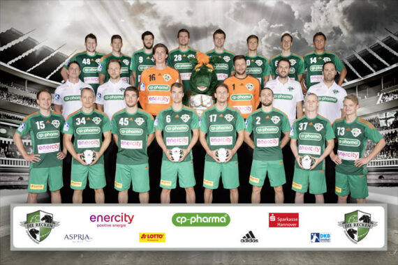 Eine Herren-Handballmannschaft in grün-weißen Trikots stellt sich in einem Stadion für ein Gruppenfoto auf. Einige Spieler halten Handbälle. Unter der Gruppe sind Sponsorenlogos zu sehen.