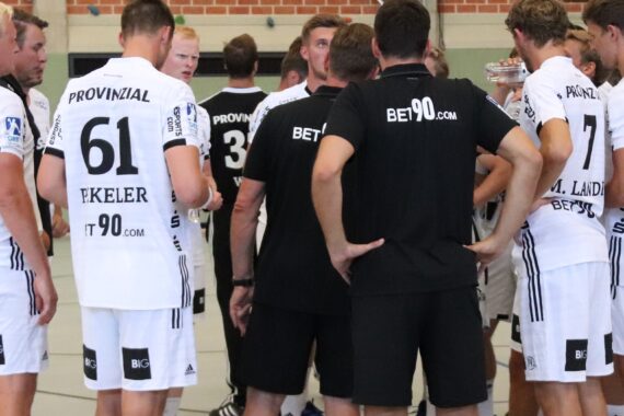 Eine Gruppe männlicher Athleten in weißen Trikots und Shorts sowie Trainer in schwarzer Kleidung versammeln sich in einer Sporthalle zu einer Mannschaftssitzung. Die meisten Trikots tragen Nummern und Sponsorenlogos. Im Hintergrund ist eine Backsteinmauer zu sehen.