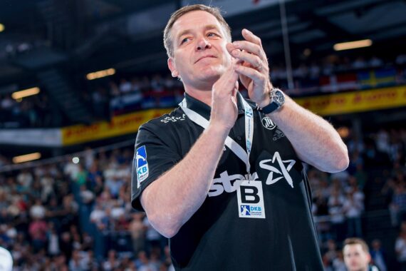 Ein Mann in einem schwarzen Sporthemd klatscht in die Hände, während er in einer mit Zuschauern gefüllten Halle steht. Er trägt ein Schlüsselband und eine Uhr und scheint die Zuschauer zu grüßen, möglicherweise bei einer Sportveranstaltung oder einer Feier.