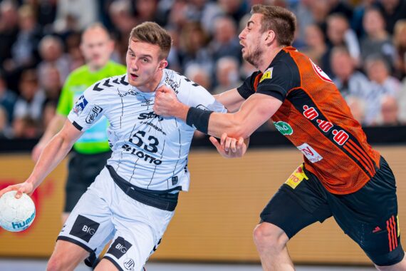 Ein Handballspieler in einem weißen Trikot versucht, an einem Verteidiger in einem schwarz-orangen Trikot vorbeizukommen, der ihn am Hals packt. Der Spieler in Weiß hält den Ball, und das Spiel wirkt intensiv. Im Hintergrund sehen Zuschauer zu.