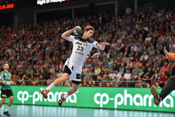 Ein Handballspieler in einem weißen Trikot springt in die Luft, um einen Schuss abzugeben, während die Zuschauer von der Tribüne aus zusehen. Das Spiel findet auf einem Hallenplatz mit leuchtend grünen Werbetafeln statt.