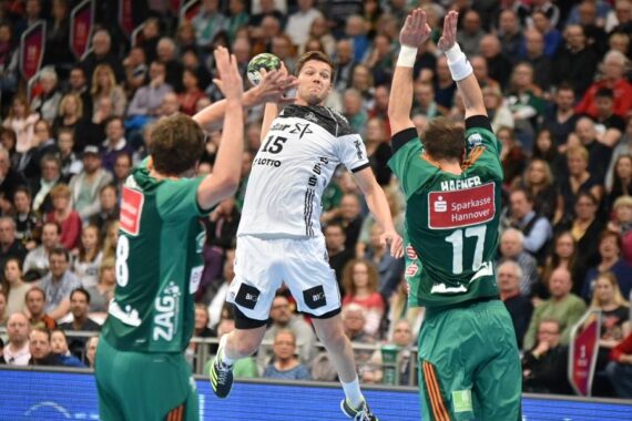 Ein Handballspieler in einem weißen Trikot springt zum Wurf, während zwei Verteidiger in grünen Trikots versuchen, ihn zu blockieren; eine Menschenmenge schaut von der Tribüne im Hintergrund zu.