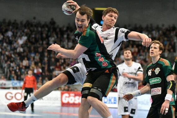 Zwei männliche Handballspieler kämpfen während eines Spiels um den Ball, wobei ein Spieler in einem grün-schwarzen Trikot springt und der andere in Weiß und Schwarz verteidigt. Das Publikum ist unscharf im Hintergrund zu sehen.