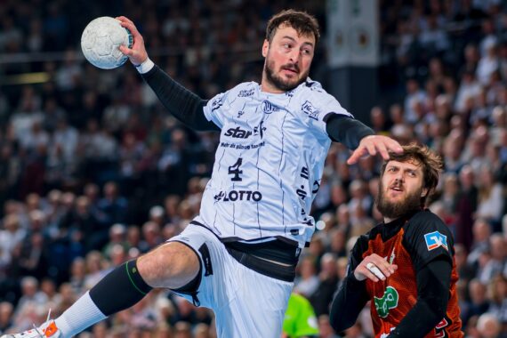 Ein Handballspieler in einem weißen Trikot springt zum Wurf, während ein Verteidiger in einem dunklen Trikot versucht, ihn zu blocken. Die Aktion findet vor einer dicht gedrängten, verschwommenen Menschenmenge statt.