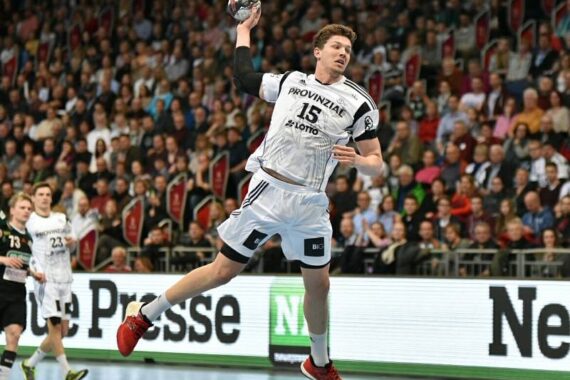 Ein männlicher Handballspieler in einem weißen Trikot springt in der Luft, um den Ball während eines Spiels zu werfen, während im Hintergrund eine Zuschauermenge von der Tribüne aus zusieht.