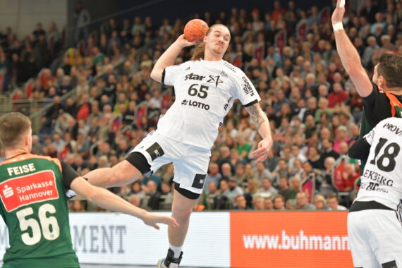 Ein Handballspieler in einem weißen Trikot bereitet sich in der Luft darauf vor, den Ball in Richtung Tor zu werfen, während die gegnerischen Spieler in Grün versuchen, ihn zu blockieren. Die überfüllte Arena verfolgt das intensive Geschehen.