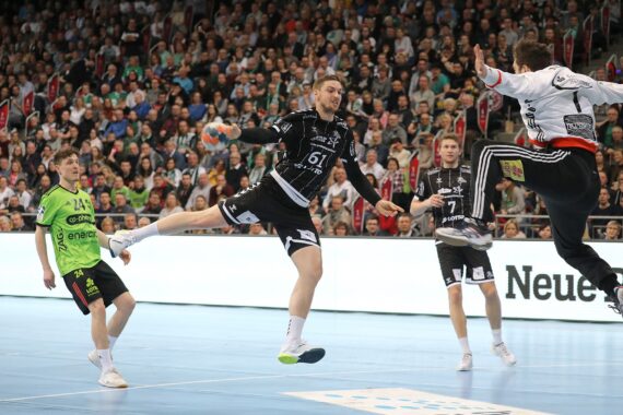 Ein Handballspieler in einem schwarzen Trikot springt in die Luft, um den Ball zu werfen, während ein Verteidiger in einem weiß-schwarzen Trikot versucht, den Ball zu blocken; andere Spieler und ein volles Publikum beobachten das Geschehen.