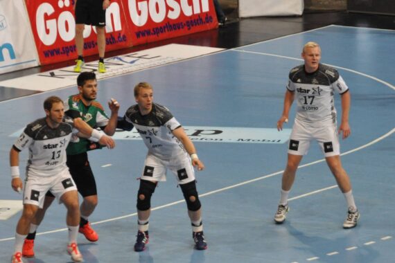 Vier männliche Handballspieler, drei in weißen Trikots und einer in einem schwarz-grünen Trikot, befinden sich während eines Spiels auf einem Hallenplatz, wobei im Hintergrund Werbebanner zu sehen sind.