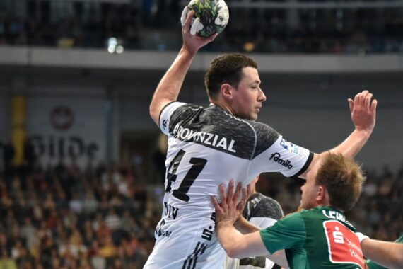 Ein Handballspieler in einem weiß-schwarzen Trikot springt, um den Ball zu werfen, während ein Verteidiger in einem grünen Trikot versucht, ihn während eines Spiels vor einer überfüllten Arena zu blockieren.