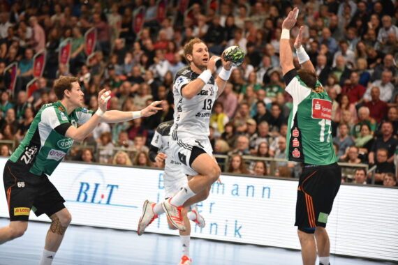 Ein Handballspieler in Weiß springt, um den Ball zu werfen, während zwei Verteidiger in Grün versuchen, ihn zu blockieren. Das Spiel findet in einer mit Zuschauern gefüllten Sporthalle statt.