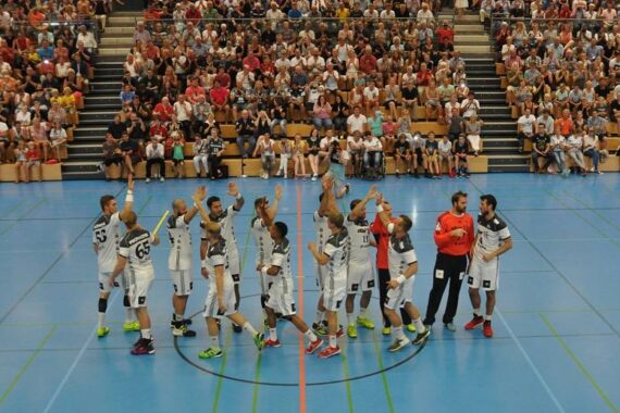 Eine Herren-Handballmannschaft in weißen Trikots gibt sich auf einem blauen Hallenplatz die Hand, während im Hintergrund eine große Zuschauermenge auf Tribünen Platz nimmt.