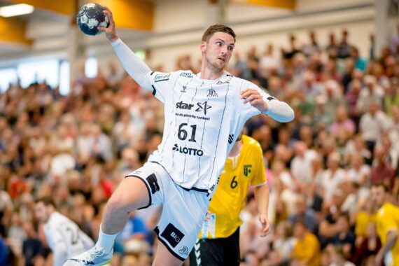 Ein männlicher Handballspieler in einem weißen Trikot bereitet sich darauf vor, den Ball während eines Spiels in die Luft zu werfen. Im Hintergrund sind eine verschwommene Menschenmenge und andere Spieler in gelben Trikots zu sehen.