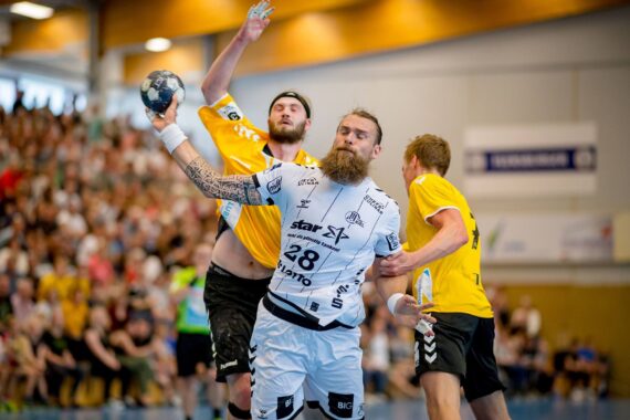 Ein bärtiger Handballspieler in einem weißen Trikot ist im Begriff, den Ball zu werfen, während er von zwei Spielern in gelben Trikots während eines intensiven Hallenspiels eng verteidigt wird, während die Zuschauer im Hintergrund zusehen.