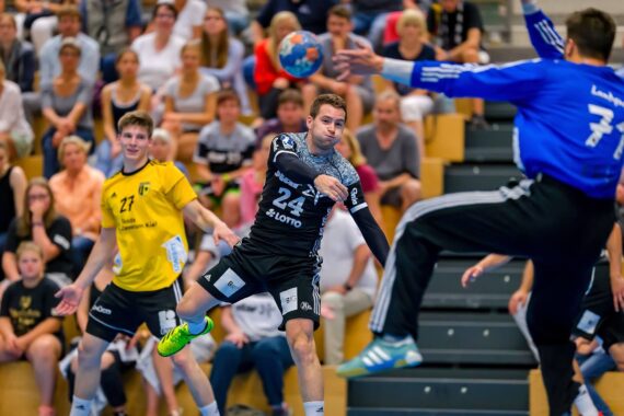Ein Handballspieler in einem schwarzen Trikot springt, um den Ball in Richtung Tor zu werfen, während der Torwart in Blau sich streckt, um ihn zu blockieren. Die Zuschauer verfolgen das Geschehen von der Tribüne aus.