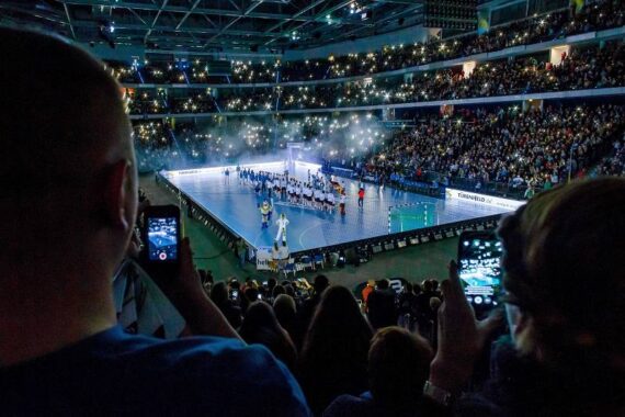 Eine große Sporthalle, gefüllt mit Zuschauern, von denen viele ihre Handys in die Höhe halten. Auf dem Spielfeld stehen die Sportler in einer Reihe unter hellen Lichtern, was eine dramatische, energiegeladene Atmosphäre schafft.