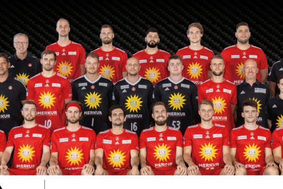 Eine Herren-Handballmannschaft posiert für ein Gruppenfoto in rot-schwarzen Trikots mit gelben Sonnenlogos. Sie stehen und sitzen in Reihen neben Trainern und Betreuern vor einem dunklen Hintergrund.