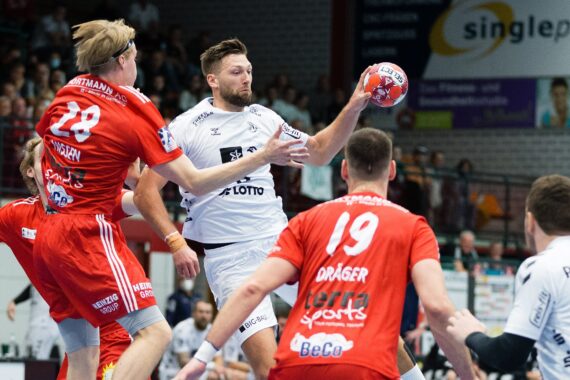 Ein männlicher Handballspieler in einem weißen Trikot springt in die Luft, um den Ball zu schießen, während drei Spieler in roten Trikots versuchen, ihn zu blockieren. Eine Menschenmenge beobachtet das intensive Spiel in einer Sporthalle.