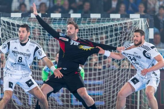 Drei männliche Handballspieler treten in der Nähe eines Tors gegeneinander an; zwei Spieler in Weiß versuchen, einen Spieler in Schwarz zu blockieren, der den Ball hält und die Trikotnummer 78 trägt. Die Intensität des Spiels ist in ihren Gesichtern zu erkennen.