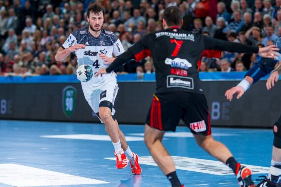Ein Handballspieler in einem weißen Trikot bereitet sich auf einen Pass oder einen Schuss vor, während er von einem Spieler in einem schwarzen Trikot in einer Halle verteidigt wird, während die Zuschauer im Hintergrund zuschauen.