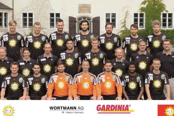 Eine Herren-Handballmannschaft posiert im Freien in drei Reihen und trägt schwarze Trikots mit einem gelben Sonnenlogo. Drei Mannschaftsmitglieder in orangefarbenen Trikots sitzen vorne in der Mitte. Sponsorenlogos erscheinen am unteren Rand. Im Hintergrund ist ein Gebäude mit Fenstern zu sehen.