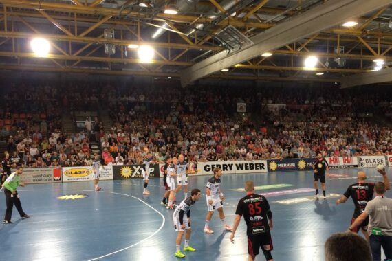 Eine überfüllte Halle, in der Mannschaften in schwarzen und weißen Trikots ein Handballspiel austragen. Die Zuschauer füllen die Tribünen, und helle Lichter erhellen das blaue Spielfeld. Trainer und Schiedsrichter stehen an der Seitenlinie.
