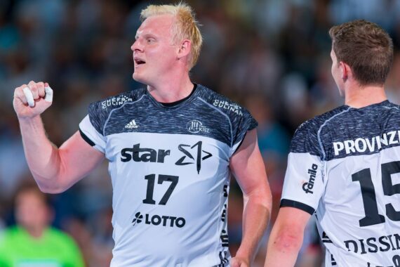 Ein männlicher Handballspieler, der ein weiß-schwarzes Trikot mit der Nummer 17 trägt, hält einen Ball in der Hand und bereitet sich auf den Wurf vor. Ein anderer Spieler mit der Nummer 15 steht neben ihm. Im Hintergrund ist eine unscharfe Menschenmenge zu sehen.