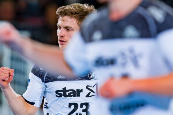 Ein Handballspieler in einem weiß-schwarzen Trikot mit der Nummer 23 jubelt mit einem Faustschlag, während ein anderer Spieler in einem ähnlichen Trikot unscharf im Vordergrund zu sehen ist.