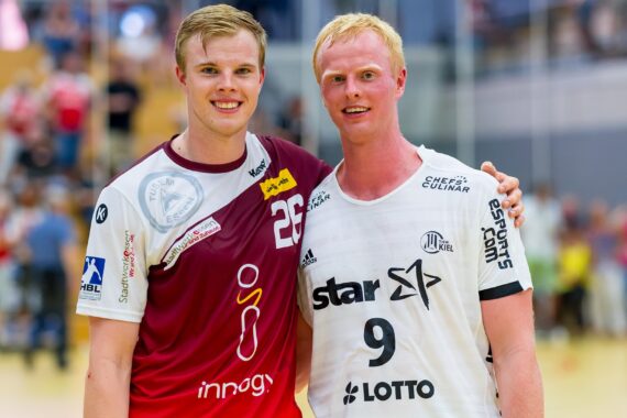 Zwei männliche Sportler in Sportuniformen stehen nebeneinander auf einem Hallenplatz und lächeln in die Kamera. Der eine trägt ein kastanienbraunes Trikot mit der Nummer 25, der andere ein weißes Trikot mit der Nummer 9. Im Hintergrund sind Zuschauer und helle Lichter zu sehen.