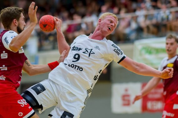 Zwei männliche Handballspieler sind in Aktion; einer im weißen Trikot (Nummer 9) versucht zu schießen oder zu passen, während er von einem Gegner im roten Trikot eng verteidigt wird. Eine unscharfe Zuschauermenge schaut im Hintergrund zu.