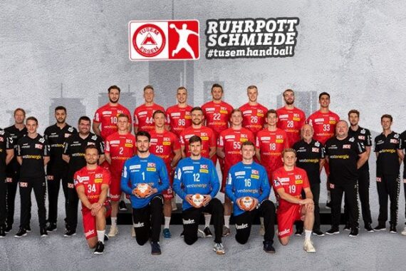 Eine Herren-Handballmannschaft posiert für ein Gruppenfoto in roten und schwarzen Trikots, wobei drei Spieler in blauen Torwarttrikots Handbälle halten. Ein grauer Hintergrund zeigt eine Stadtsilhouette und die Aufschrift RUHRPOTTSCHMIEDE tusen.handball.