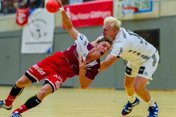 Zwei männliche Handballspieler liefern sich einen intensiven Wettkampf; der eine in Rot und Weiß wird von einem Gegner in Weiß zu Boden gezogen, beide sind aus dem Gleichgewicht und greifen nach dem Ball auf einem Hallenplatz. Im Hintergrund sind Zuschauer und Fahnen zu sehen.