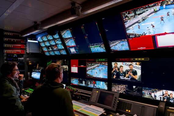 Zwei Personen sitzen an einer Schalttafel in einem Fernsehstudio und überwachen mehrere Bildschirme, auf denen Live-Sportaufnahmen und Kameraperspektiven zu sehen sind, darunter ein Volleyballspiel und Kommentatoren. Der Raum ist voll mit Geräten und leuchtenden Monitoren.