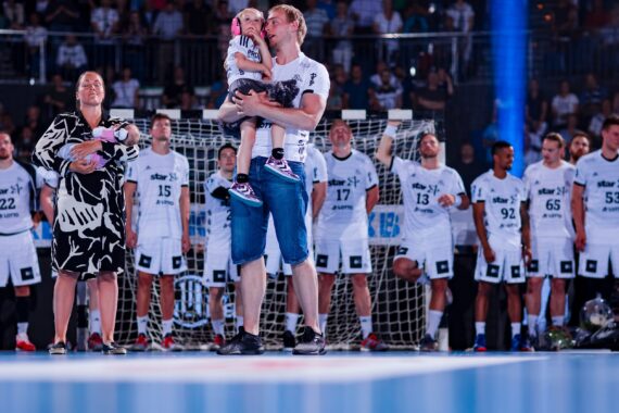 Ein Mann mit einem Kind im Arm steht auf einem Handballfeld, daneben hält eine Frau ein Baby. Hinter ihnen steht eine Mannschaft in weißen Trikots vor einem Tor, im Hintergrund sind Zuschauer zu sehen.