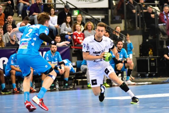 Ein Handballspieler in einem weißen Trikot läuft mit dem Ball, während er von einem Spieler in einem blauen Trikot während eines Spiels verteidigt wird, während die Zuschauer auf der Tribüne im Hintergrund zusehen.