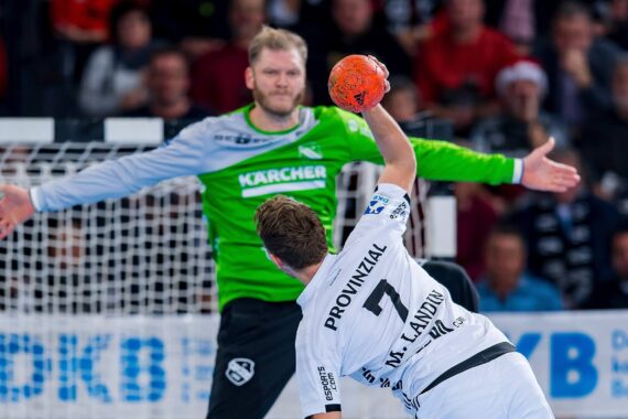 Ein Handballspieler in einem weißen Trikot bereitet sich darauf vor, auf das Tor zu schießen, während der Torwart in Grün mit weit geöffneten Armen dasteht, bereit zum Abwehren. Das Spiel findet in einer Halle statt, mit einer unscharfen Menschenmenge im Hintergrund.