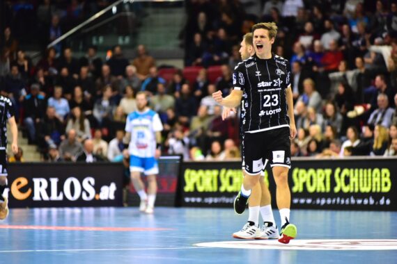 Ein männlicher Handballspieler, der ein schwarzes Trikot mit der Nummer 23 trägt, jubelt während eines Spiels auf einem Hallenplatz. Im Hintergrund sitzen Zuschauer, und andere Spieler sind auf dem Spielfeld zu sehen.