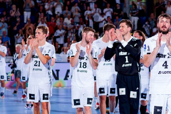 Eine Gruppe von männlichen Handballspielern in weißen Uniformen, von denen einige klatschen, läuft gemeinsam auf einem Hallenplatz mit einer jubelnden Menge im Hintergrund.